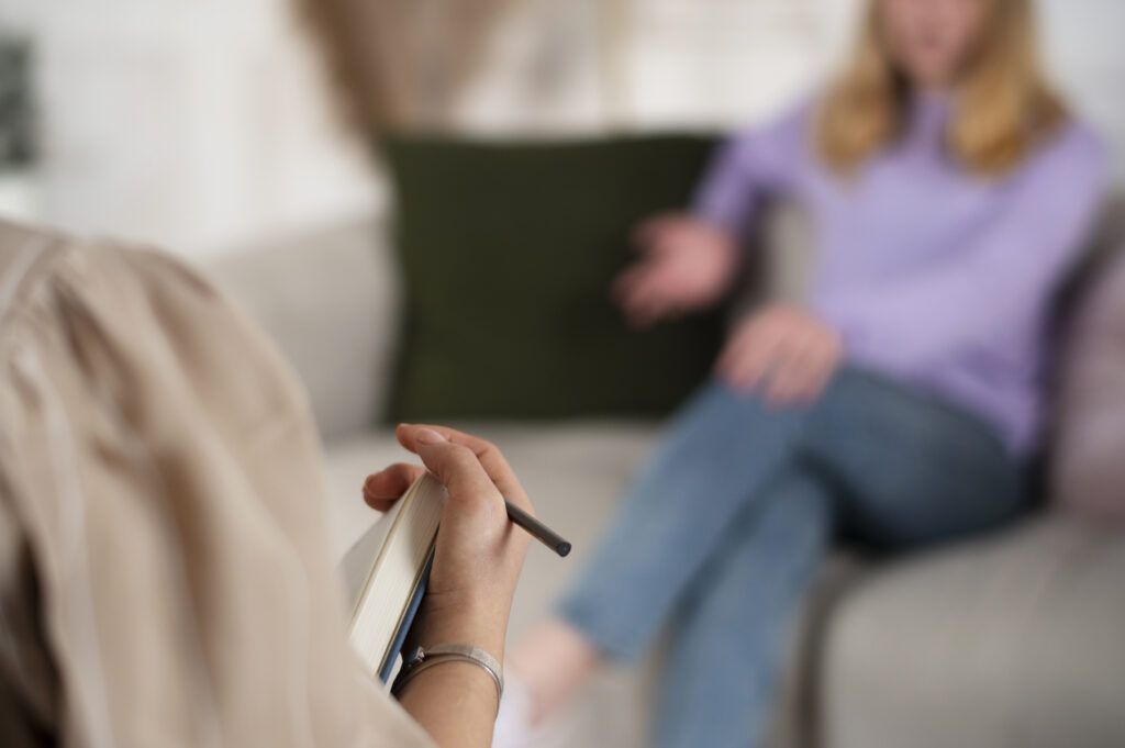 
A imagem mostra a perspectiva de um terapeuta, com a mão segurando um caderno e uma caneta, enquanto ouve um paciente. O paciente, uma pessoa com suéter roxo e calça jeans, está sentado em um sofá no fundo, fora de foco. A cena sugere uma sessão de terapia, focando na escuta e anotações do profissional.