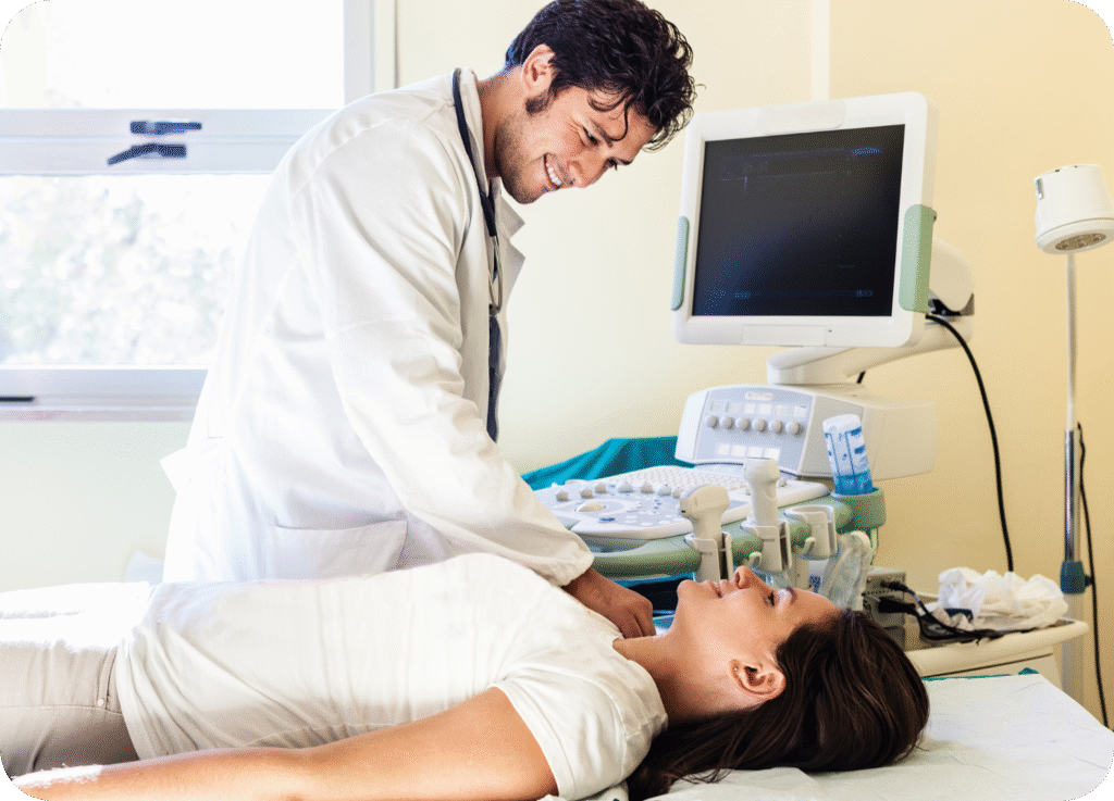 
Um médico sorridente, vestindo um jaleco branco e estetoscópio, realiza um exame de ultrassonografia em uma paciente feminina. O médico está aplicando o transdutor na região do pescoço da paciente, sugerindo um ultrassom de tireoide ou de carótidas. A paciente está deitada confortavelmente em uma maca, vestindo uma camiseta branca, enquanto o monitor do equipamento de ultrassom exibe as imagens em tempo real ao lado. A cena transmite um ambiente clínico de atendimento atencioso.