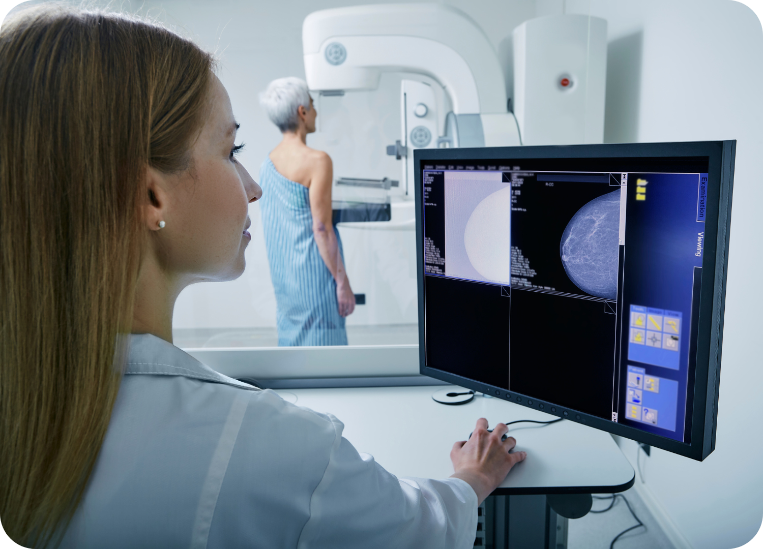 Uma profissional de saúde, possivelmente uma técnica em radiologia ou médica, está sentada de perfil em frente a um monitor de computador. Ela está vestida com um jaleco branco e observa atentamente a tela, onde é exibida uma imagem de mamografia ou um exame de imagem das mamas. Ao fundo, ligeiramente desfocada, uma paciente idosa (com cabelos grisalhos) está em pé, de costas para a câmera e vestida com uma bata hospitalar azul, posicionada dentro de um aparelho de mamografia. A cena sugere um ambiente de clínica de exames de imagem focado na saúde da mulher e no diagnóstico por imagem.