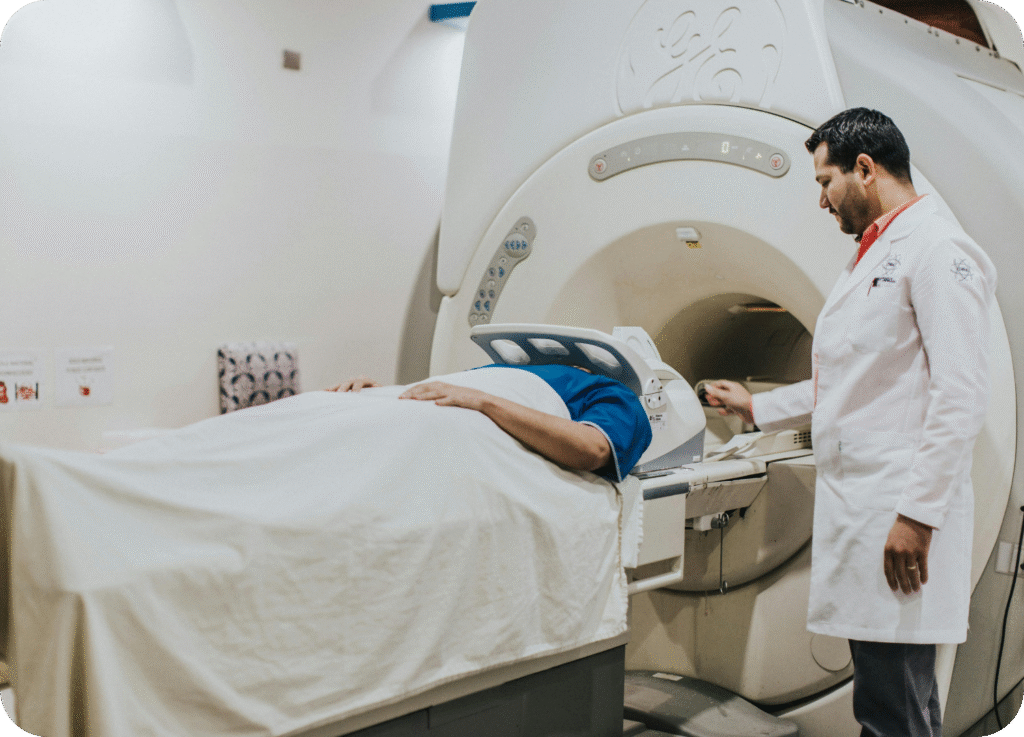
Um técnico ou médico radiologista (homem de jaleco branco) supervisiona um paciente deitado em uma maca, prestes a iniciar um exame de Ressonância Magnética (RM) ou Tomografia Computadorizada (TC). O paciente está coberto por um lençol branco, com a parte superior do corpo voltada para a abertura do grande equipamento de imagem (o gantry). O profissional de saúde está concentrado, interagindo com os comandos do aparelho, o que sugere o início de um procedimento diagnóstico detalhado.