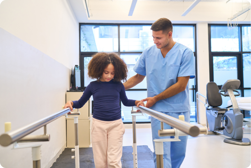 Um profissional de saúde de pediatria jovem, vestindo um uniforme médico azul claro, auxilia uma menina com cabelo cacheado volumoso em um exercício de caminhada. A menina segura-se em barras paralelas de metal para se equilibrar enquanto caminha sobre um tatame preto. O ambiente é uma clínica de reabilitação ou fisioterapia bem iluminada, com grandes janelas ao fundo e equipamentos de exercício visíveis.
