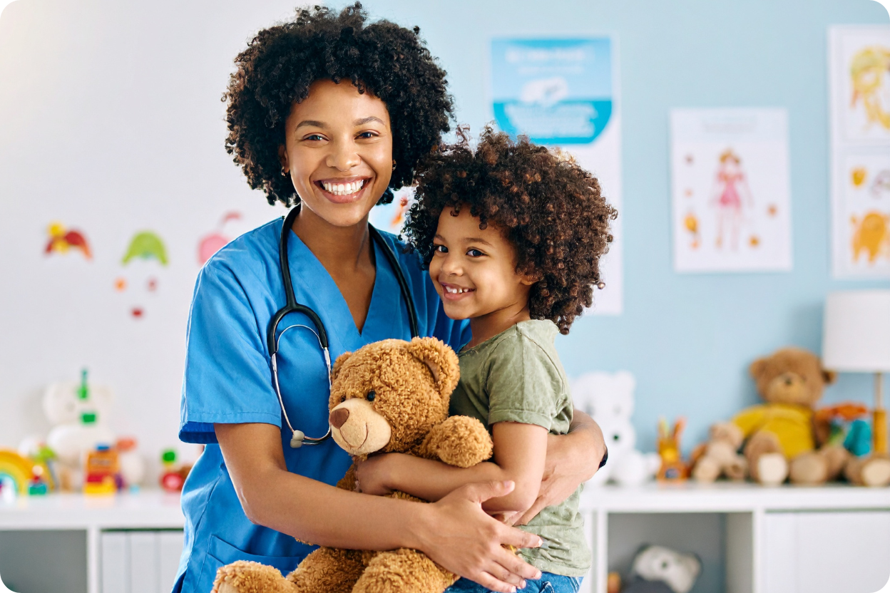 Uma mulher negra sorridente, vestindo um uniforme médico azul (scrub) e com um estetoscópio no pescoço, abraça carinhosamente uma criança negra, que segura um urso de pelúcia marrom. Ambas sorriem para a câmera em um ambiente que parece ser um consultório pediátrico alegre, com brinquedos e desenhos coloridos ao fundo.