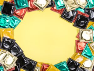 Diversos pacotes coloridos de preservativos dispostos em círculo sobre um fundo amarelo vibrante, com um espaço vazio redondo no centro para texto.