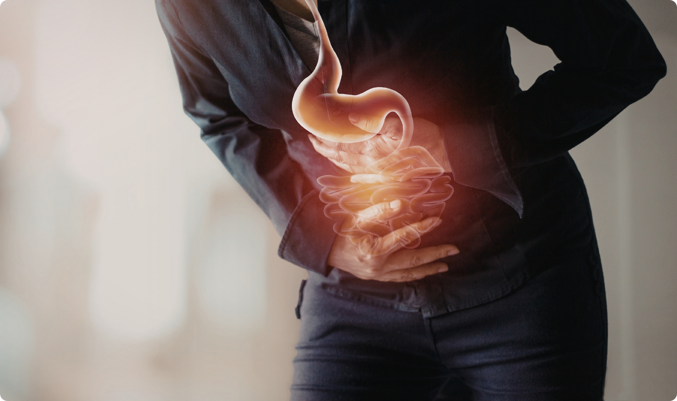 Imagem com efeito de sobreposição mostrando uma pessoa vestida com roupas escuras, inclinada para frente e pressionando a região abdominal com as mãos. Sobre o corpo, há uma representação gráfica iluminada do sistema digestório, destacando o estômago e os intestinos com um brilho alaranjado, simbolizando dor ou inflamação.