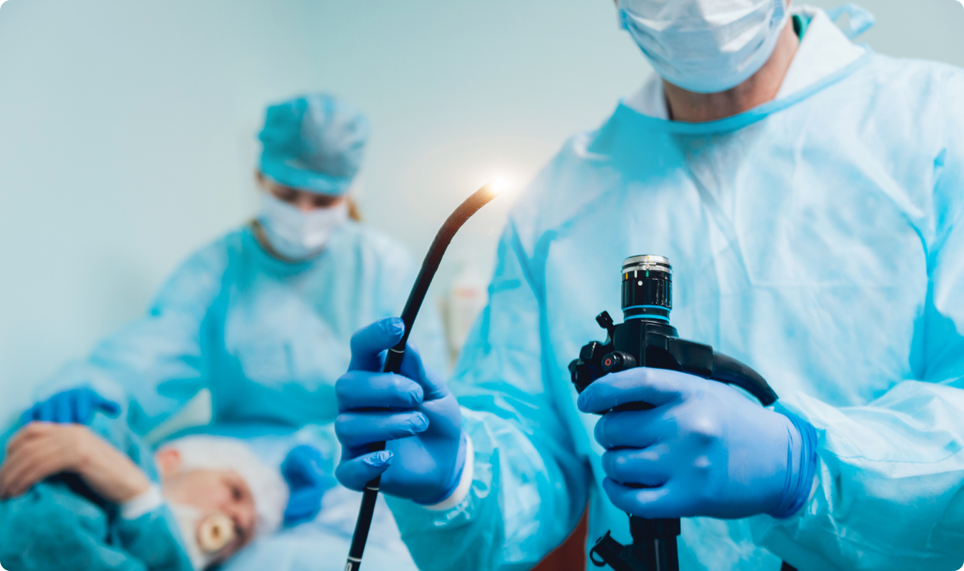 A imagem mostra um procedimento de endoscopia sendo realizado em um paciente deitado em uma maca em um ambiente de hospital. Um profissional de saúde, usando luvas e máscara, segura o endoscópio, um instrumento longo e fino com uma câmera e luz, enquanto o endoscópio é inserido na boca do paciente. O paciente parece estar acordado e olhando para o procedimento. O monitor de endoscopia exibe imagens das partes internas do trato digestivo do paciente, visíveis ao médico e à equipe que o auxilia. A sala está limpa e organizada, com equipamentos médicos ao redor.